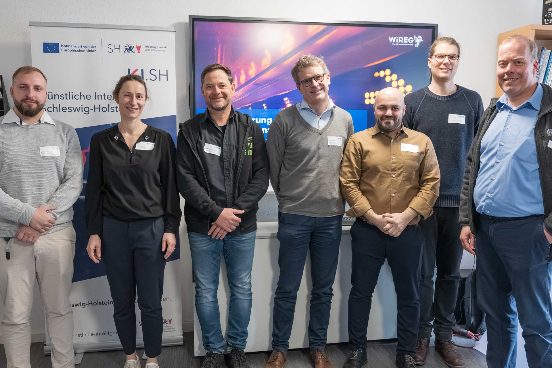 Die Workshop-leitenden Personen stehen vor einem KI.SH-Banner und einer Leinwand, auf der das WiREG-Logo zu sehen ist